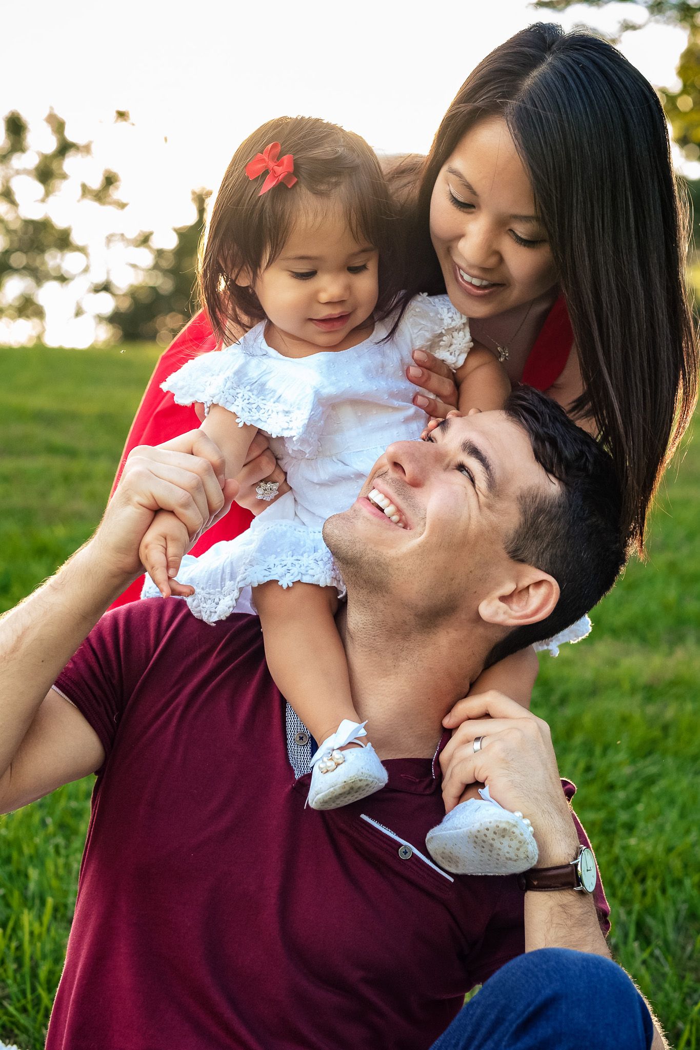 Joyful family sitting on grass at sunset, parents smiling with their baby daughter in a white dress.