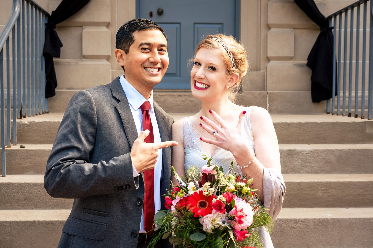 Newlyweds smile and show off wedding ring on courthouse steps with colorful bridal bouquet.