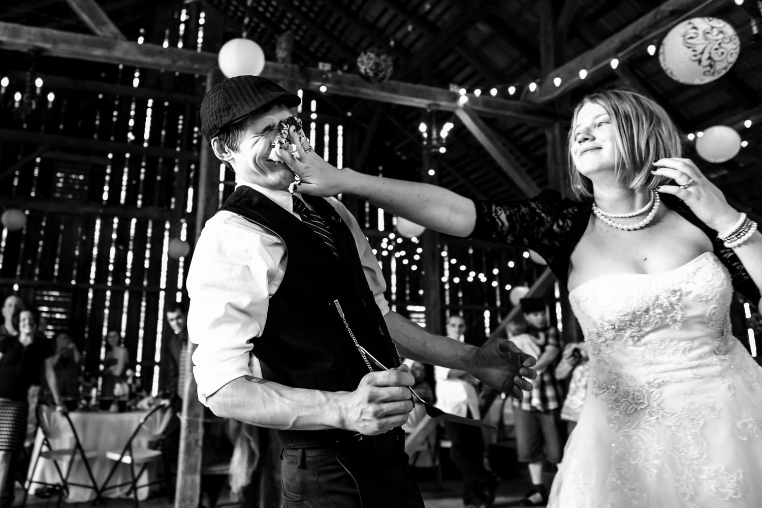 Bride and groom share a joyful dance inside a rustic barn decorated with string lights.
