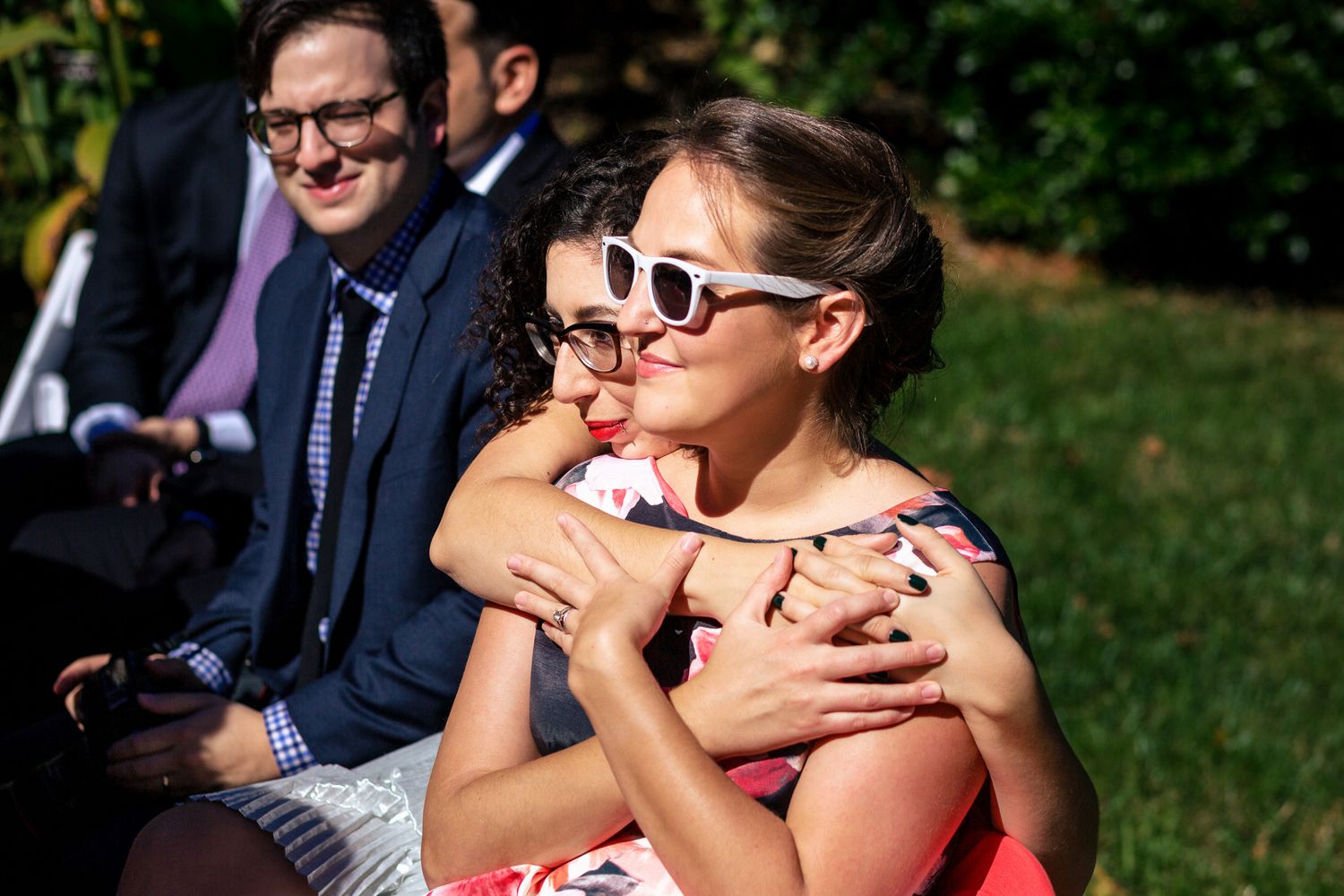 Two women embrace warmly while watching outdoor wedding ceremony on sunny day.