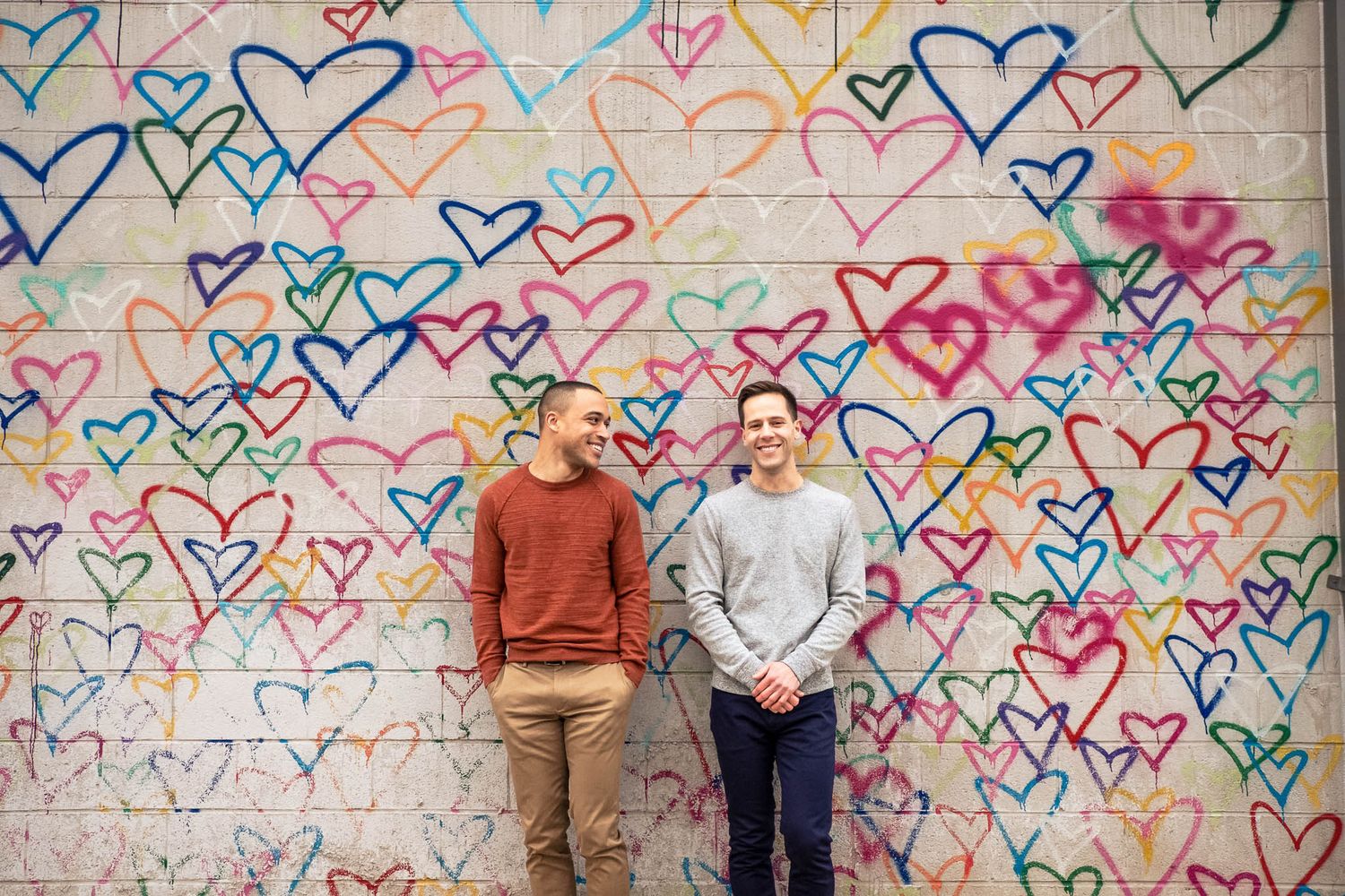 Two men smiling together in front of a colorful wall covered in painted rainbow hearts.
