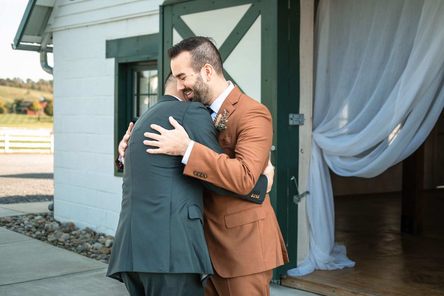 Two grooms embrace warmly during their first look at an elegant barn wedding venue.