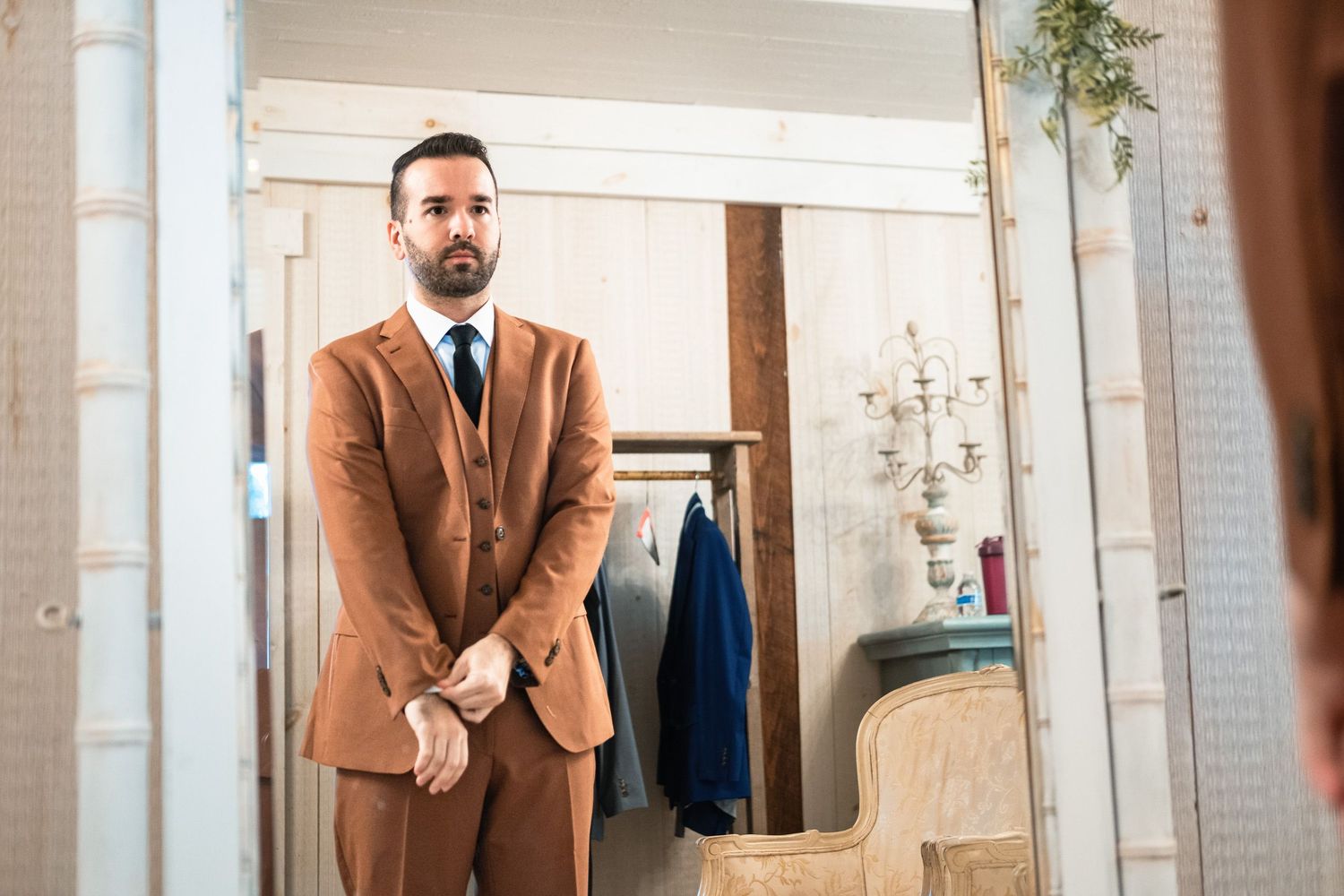 Groom in stylish brown three-piece suit adjusting jacket while looking in mirror.