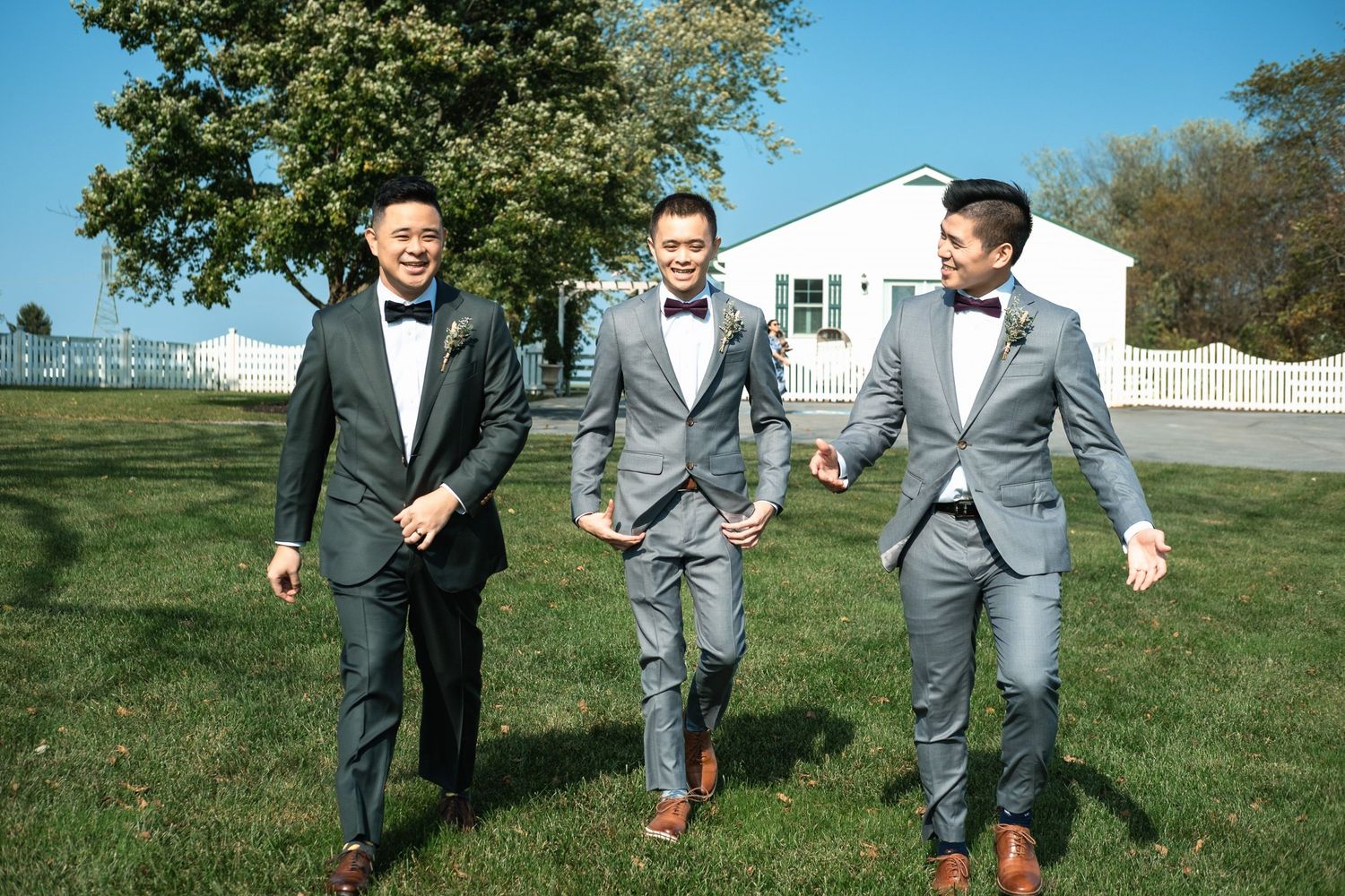 Three groomsmen in matching gray suits and bow ties walking together on sunny wedding day.
