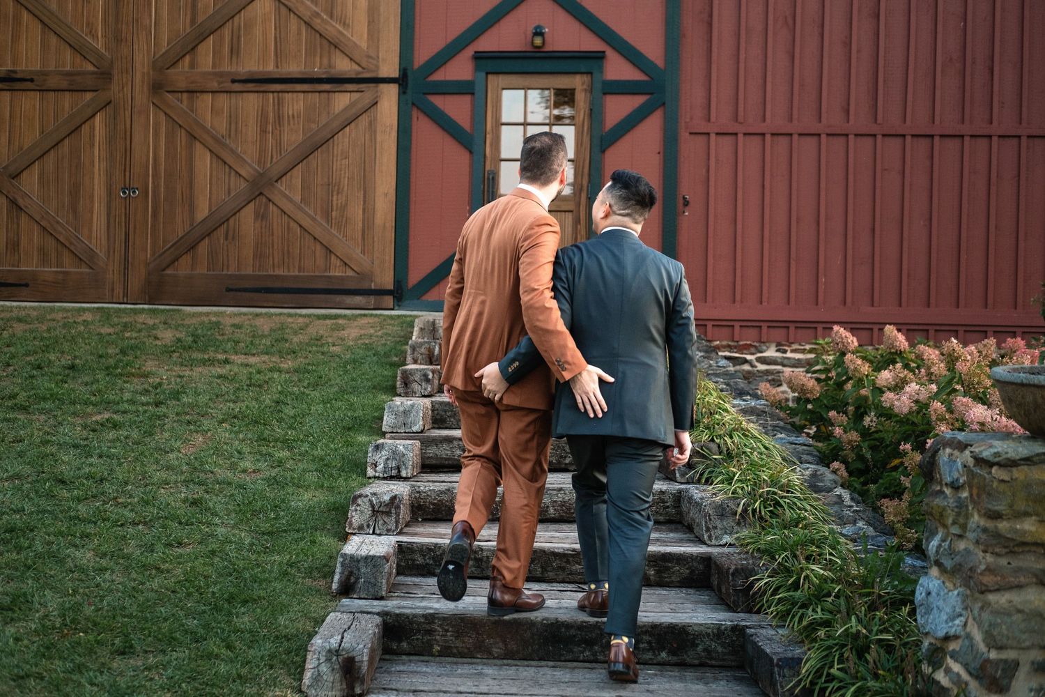 Two grooms in suits walk up stone steps toward a red barn wedding venue together.