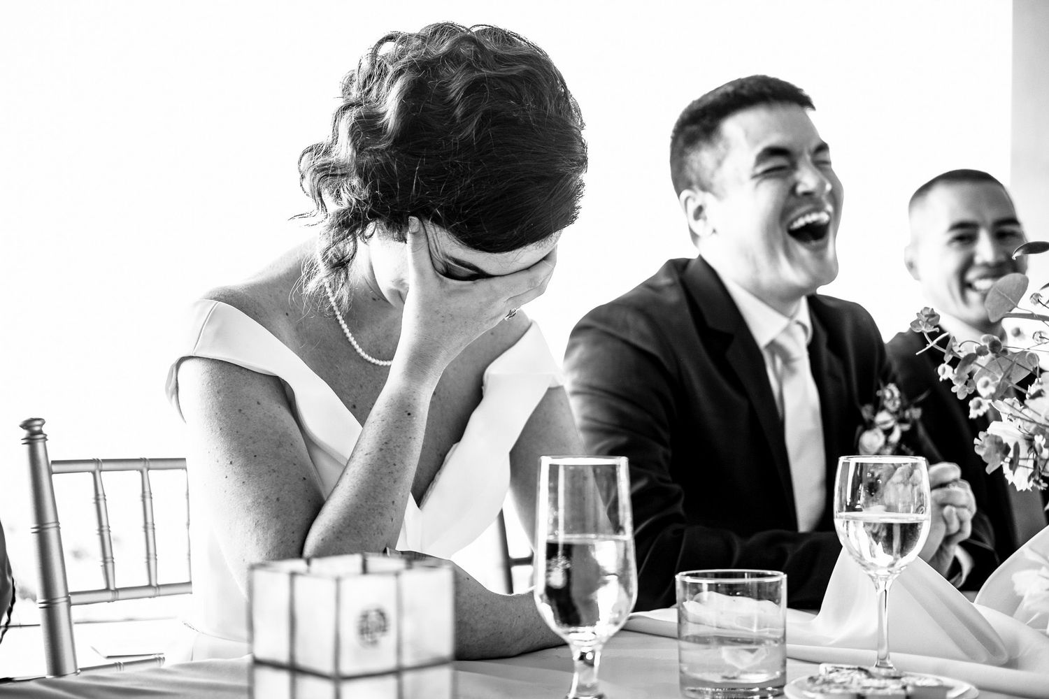Bride laughing and covering her face while groom smiles during wedding reception speeches.