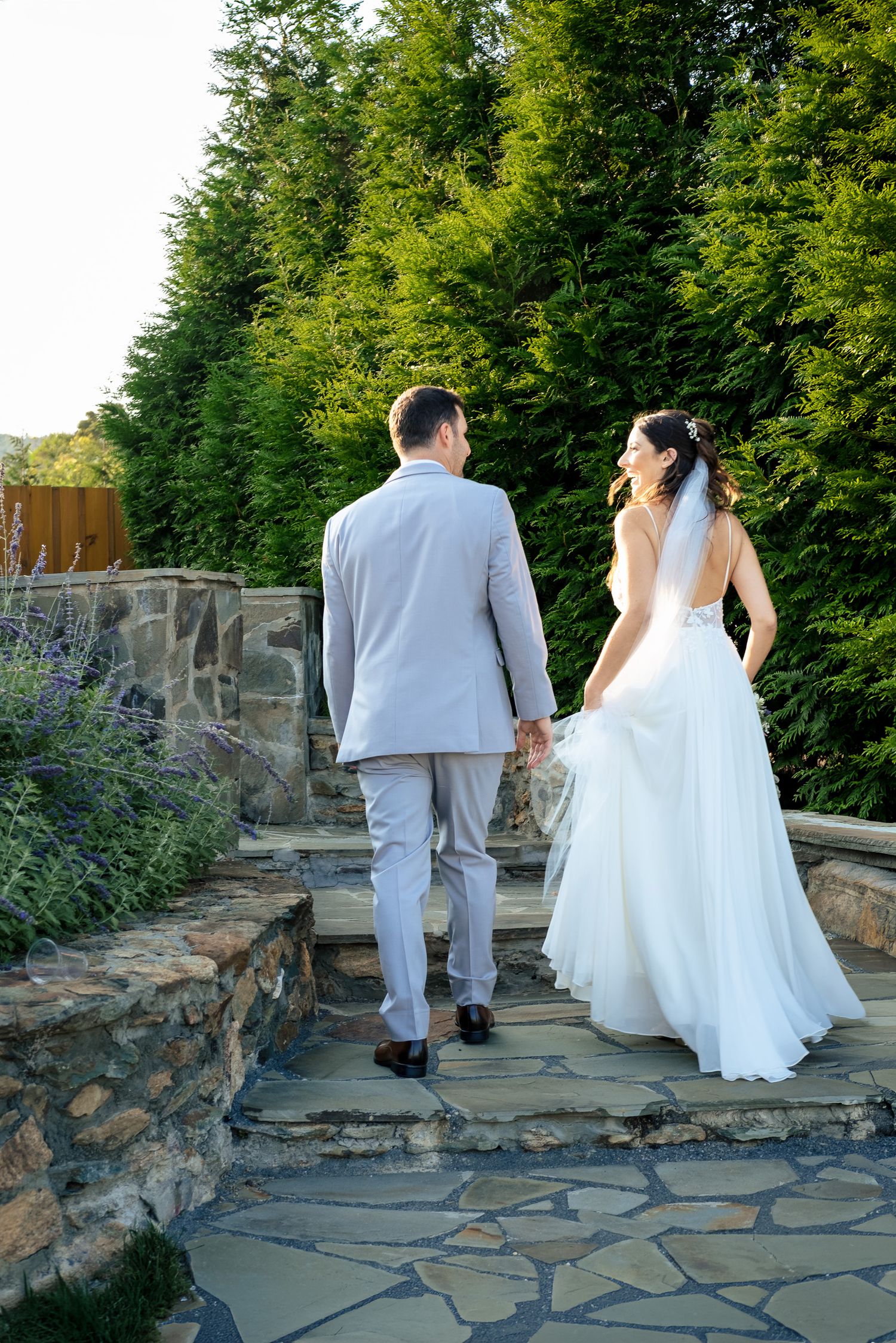 Bride and groom walk hand in hand up stone steps surrounded by lush green trees and garden landscaping.