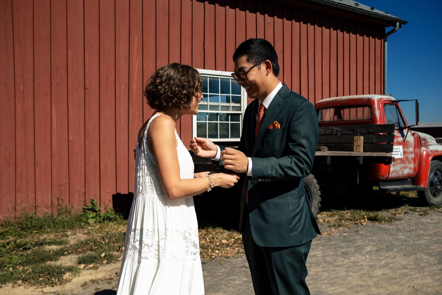 A groom adjusts his bride's jewelry outside a rustic red barn on their wedding day.