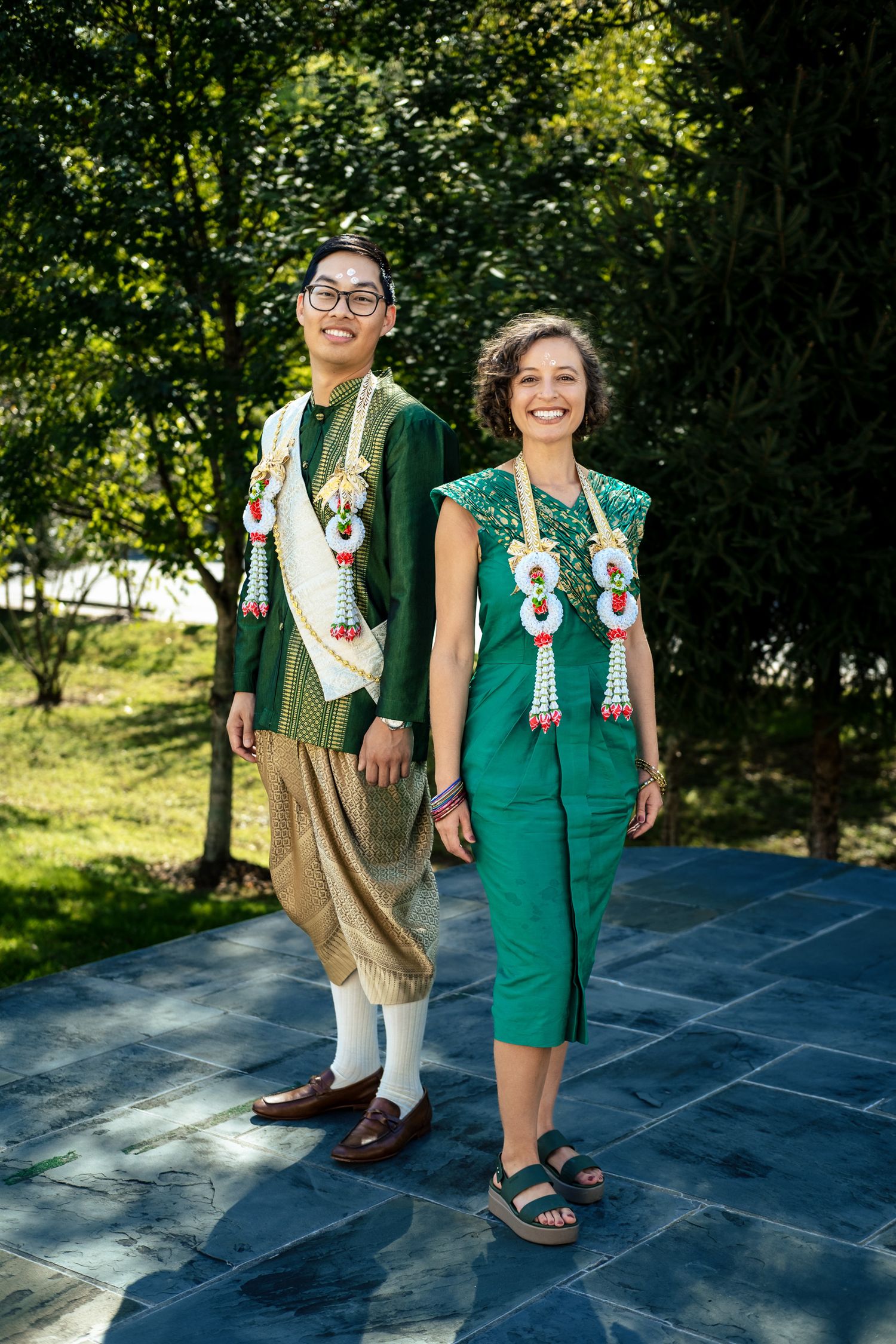 Couple wearing traditional Thai green wedding outfits with floral garlands standing outdoors.