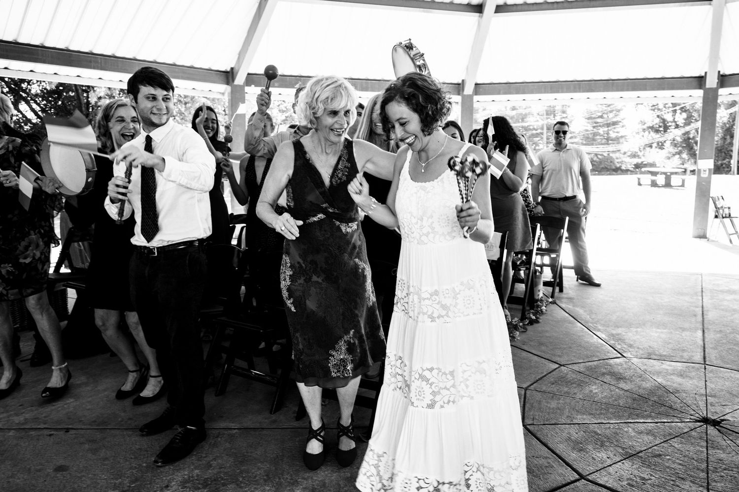 A bride in a white lace gown is escorted by two guests in black and white wedding photo.