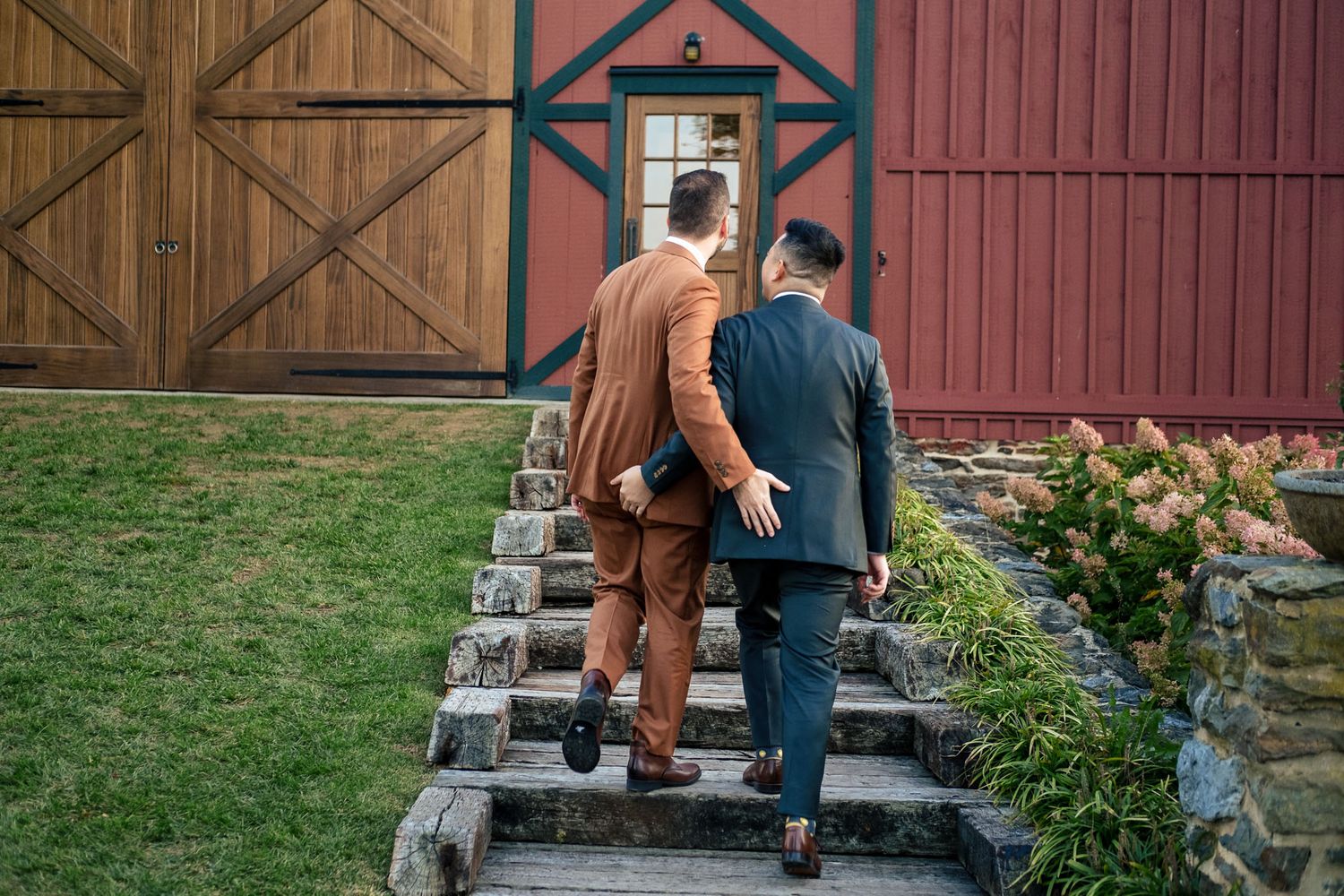 Two grooms in suits walk up stone steps toward a rustic red barn wedding venue.