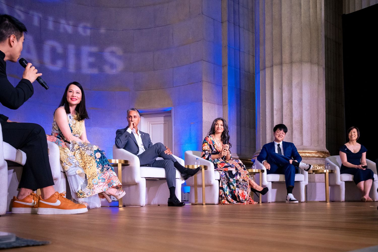 Panel of six speakers seated on stage at a formal conference event with decorative columns in the background.