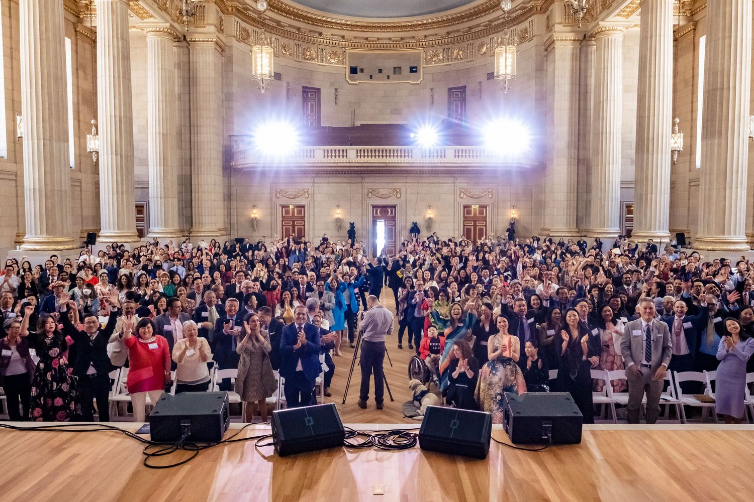Large crowd gathered in an ornate grand hall with bright stage lights during a formal group event or ceremony.