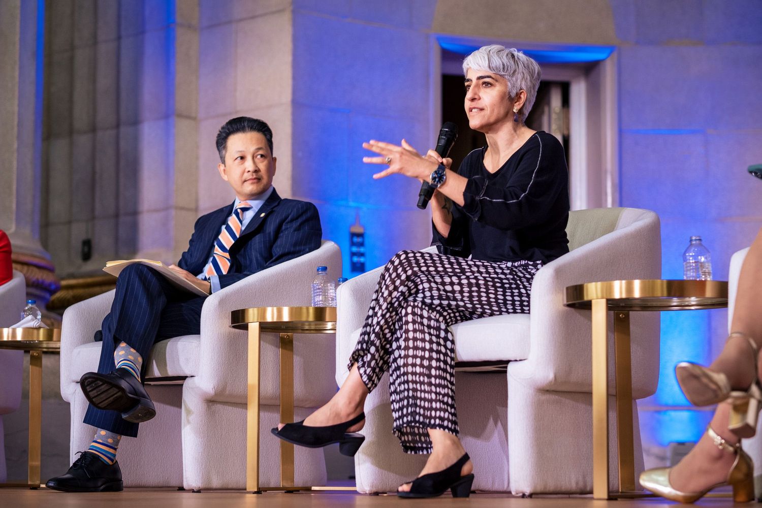 Two panelists speak on stage at a professional conference, woman with gray hair gestures while holding microphone.