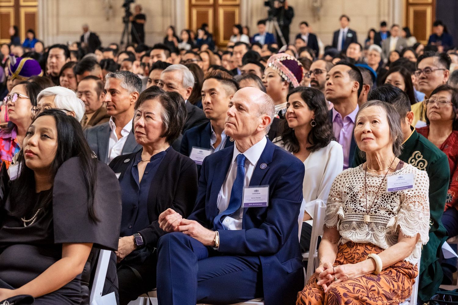 Diverse crowd of attendees seated at a formal multicultural ceremony or civic event in an elegant hall.
