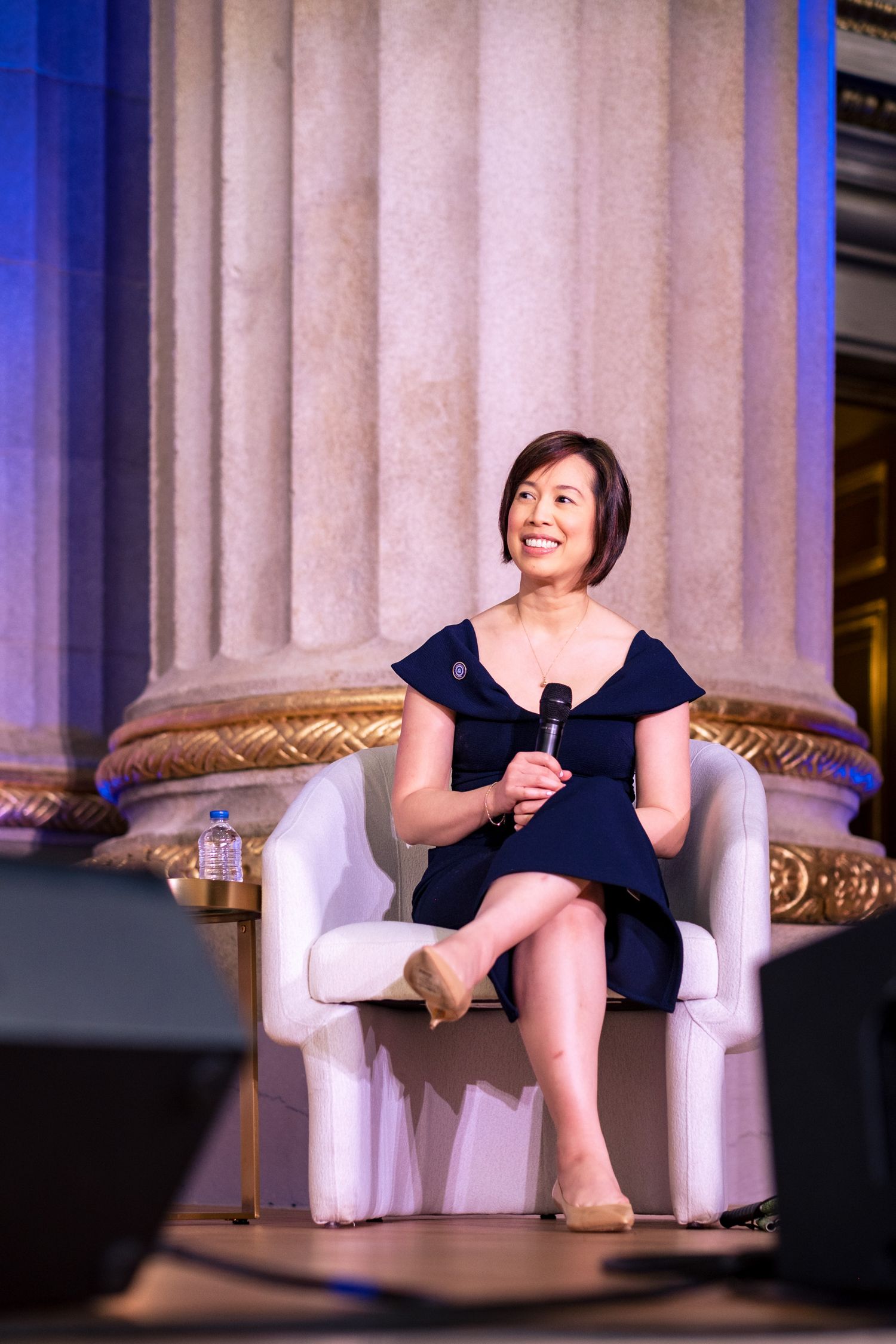 Woman in navy dress sits in white chair holding microphone, speaking at elegant event with marble columns.