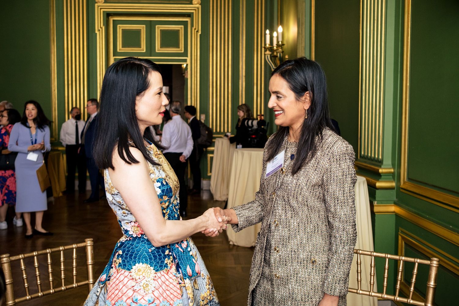 Two women shaking hands and networking at a formal event in an elegant green and gold ballroom.