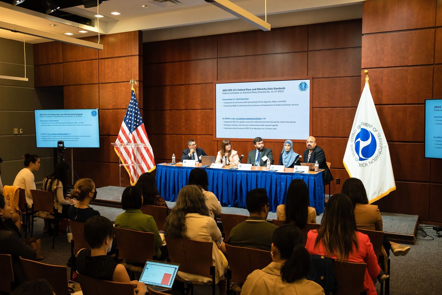 Panel discussion at a formal conference with speakers at a blue table, US flag, and audience members seated in rows.