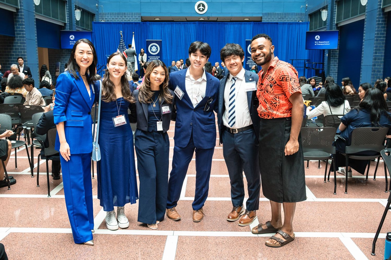 Group of seven diverse students posing together at a formal academic event in a blue-themed hall.