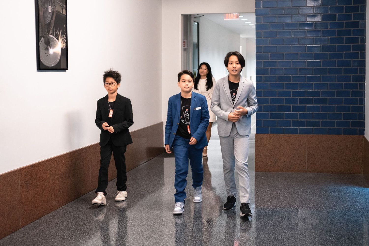 Four people walking down a hallway with blue tile walls, dressed in stylish suits and casual outfits.