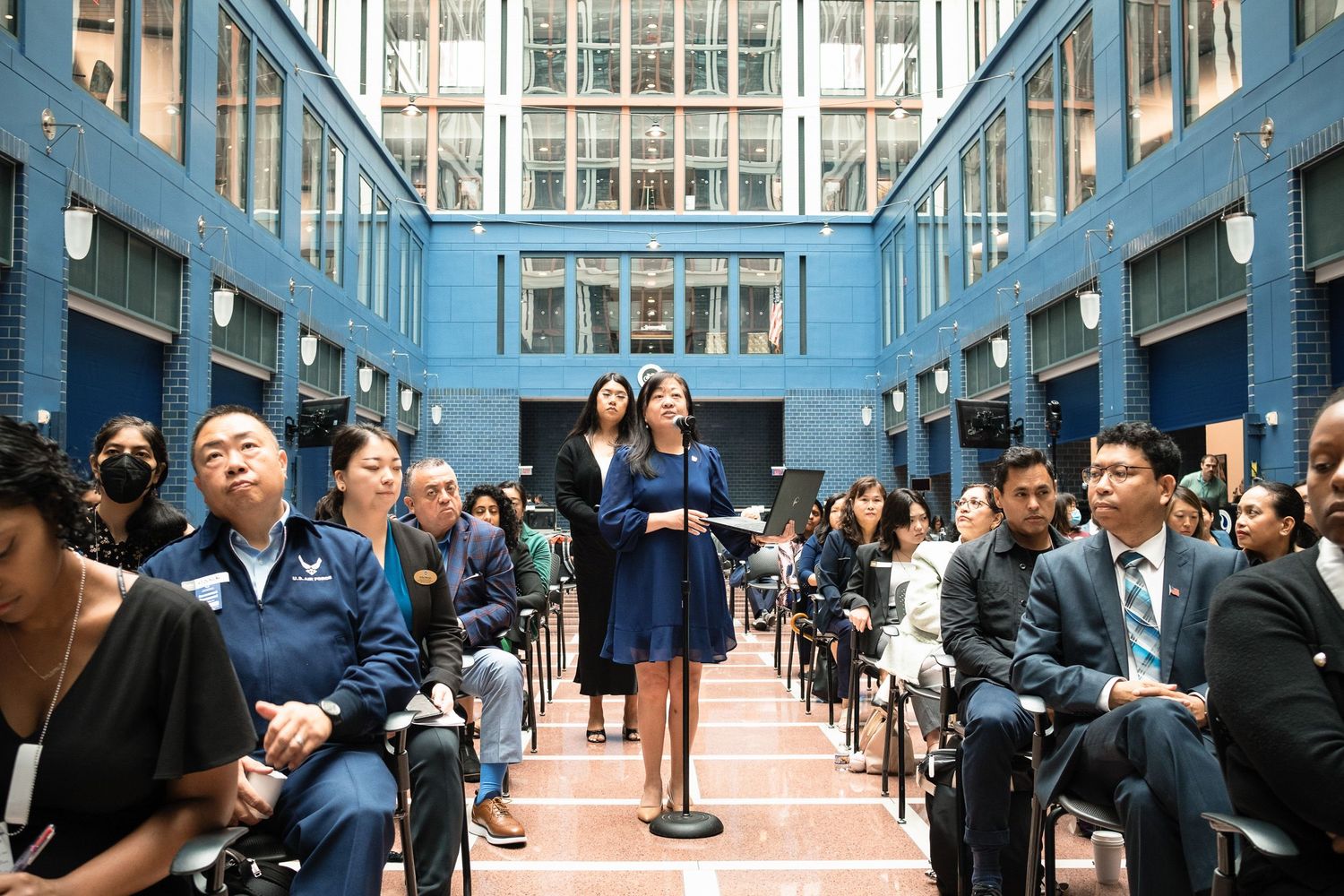 Woman speaks at podium during formal indoor ceremony in blue-tiled atrium with seated audience.