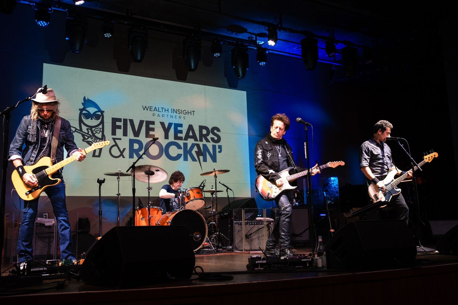 Rock band performs on stage at Five Years & Rockin anniversary event with guitarist, drummer, and bassist under blue spotlight.