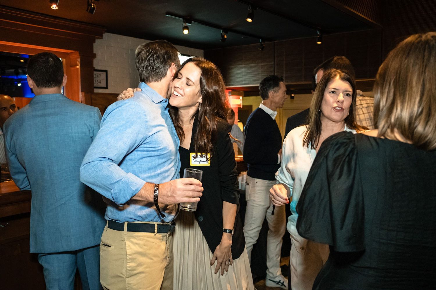 Guests mingling and socializing at an indoor networking event, smiling and holding drinks in a dimly lit venue.
