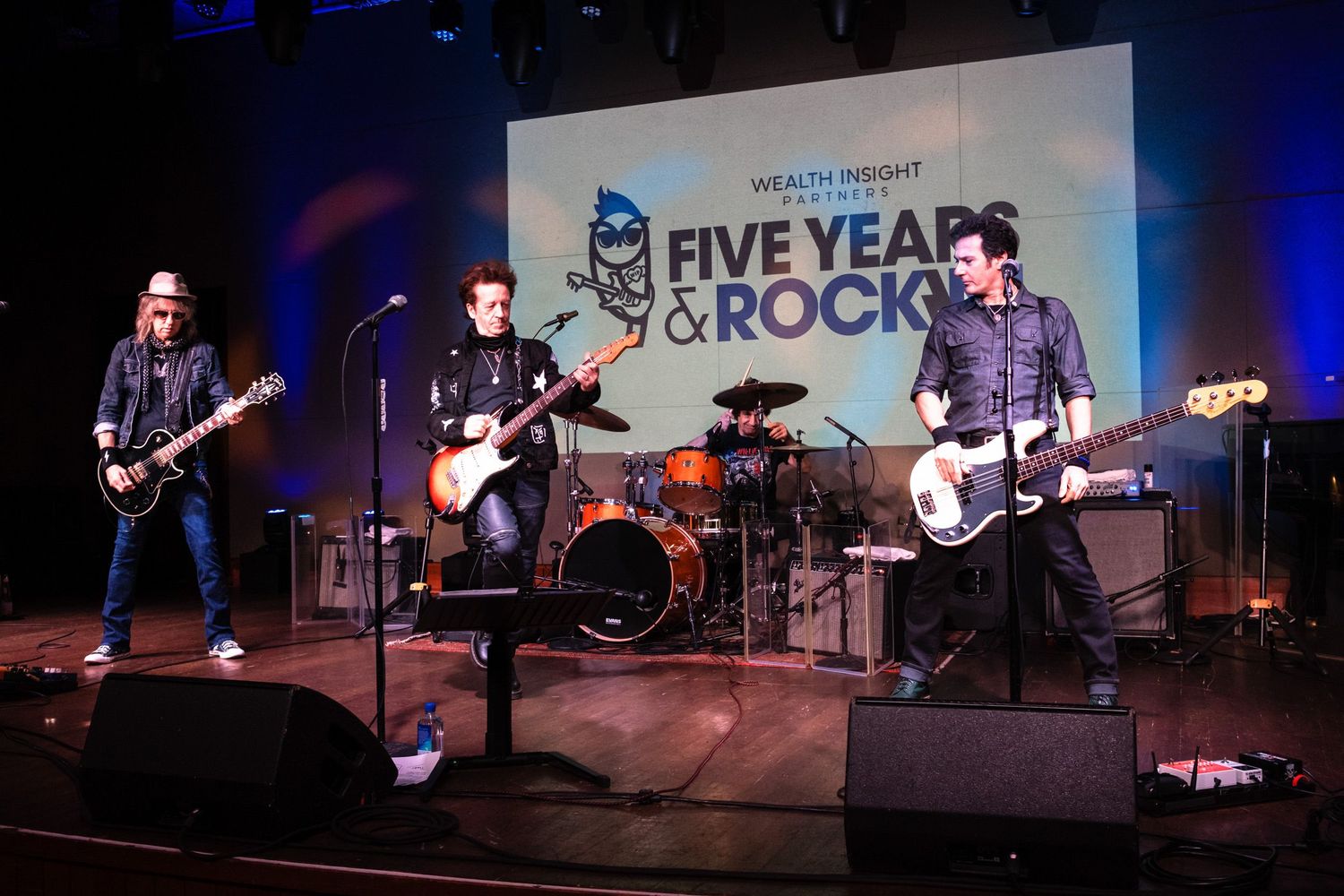 Rock band performing on stage at Wealth Insight Five Years & Rock anniversary event with guitars and drums.