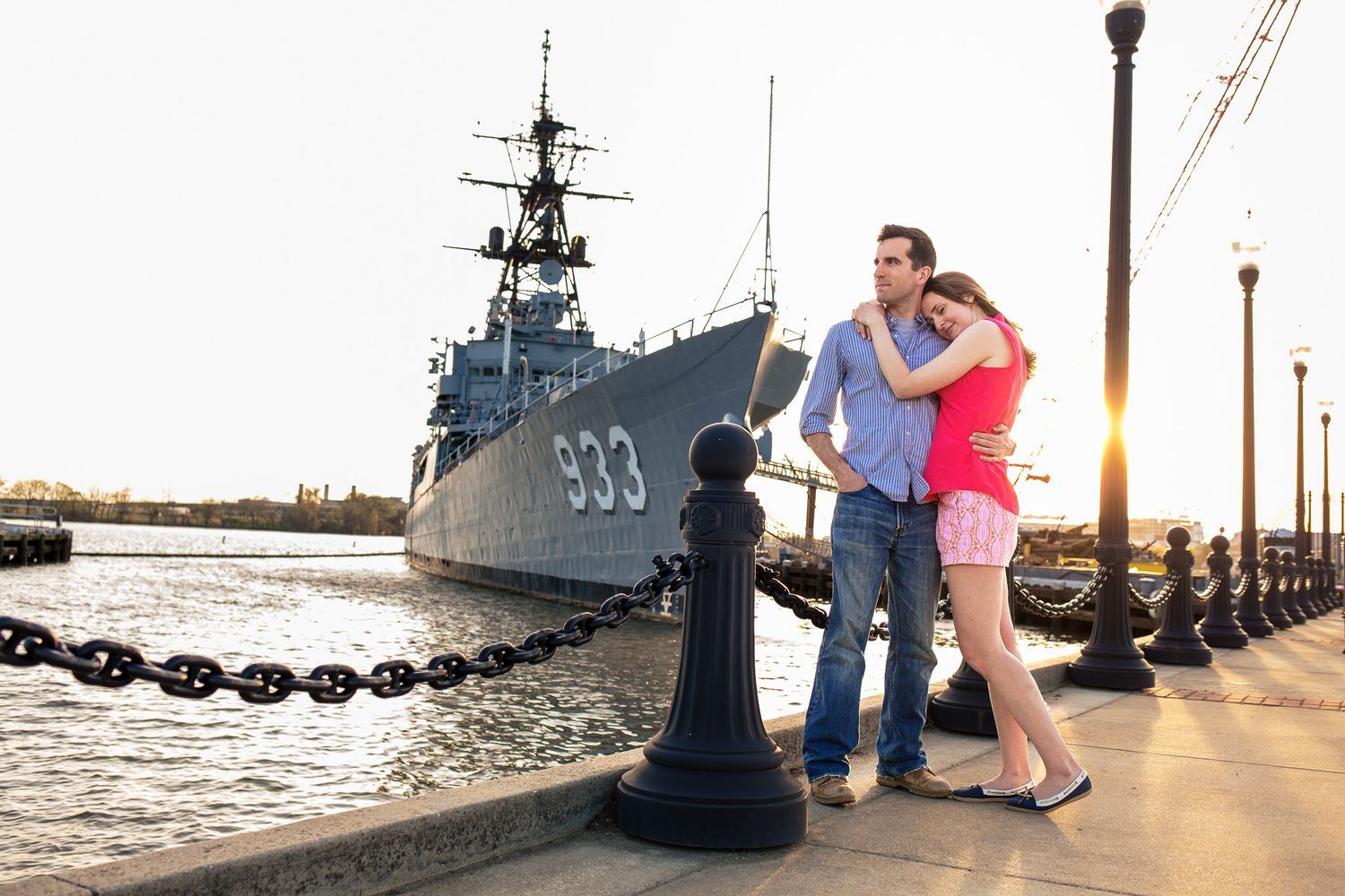 Couple embracing at sunset on a waterfront promenade with a naval destroyer ship behind them.