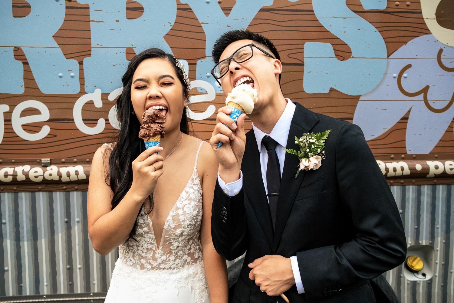 Joyful newlyweds in wedding attire laugh while eating ice cream cones outside an ice cream shop.