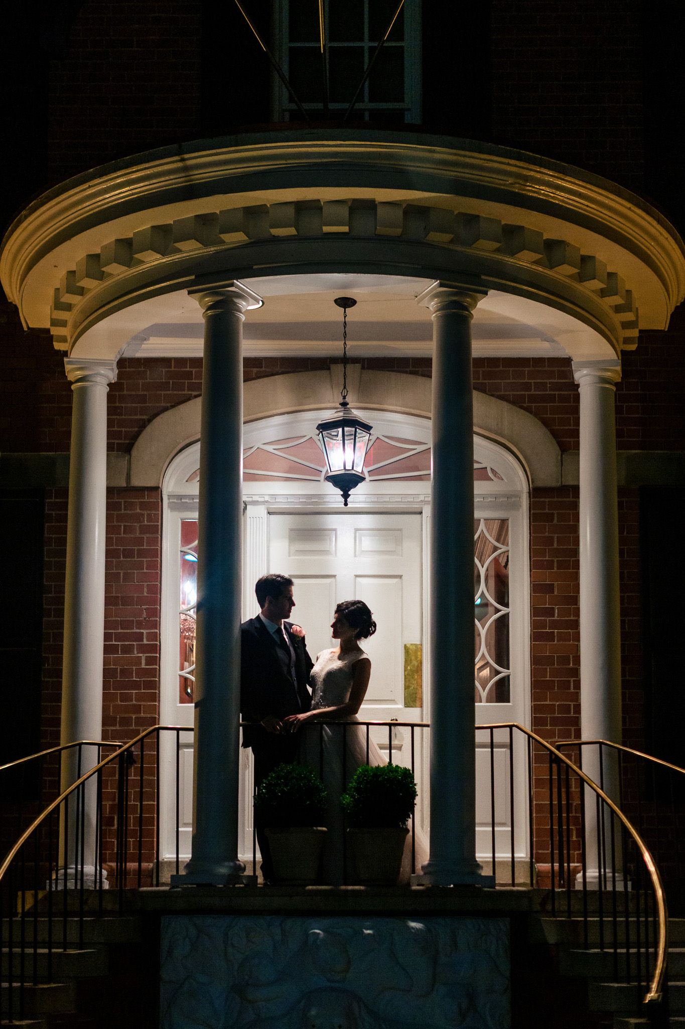 Bride and groom share a romantic silhouette moment on a lit colonial brick building porch at night.