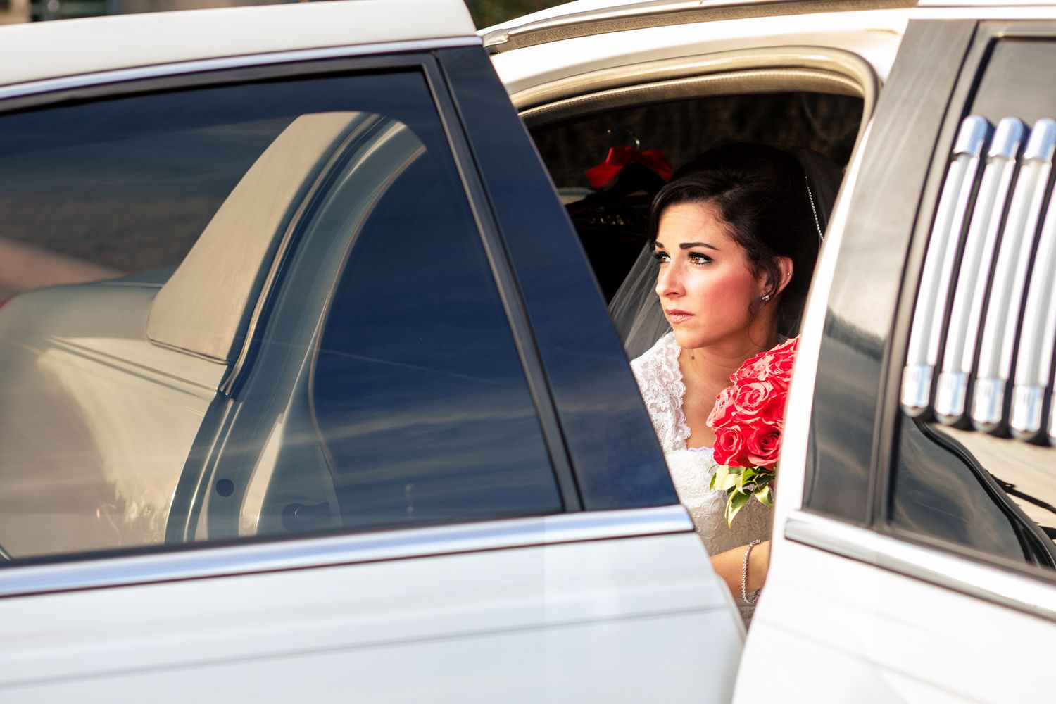 Bride in white dress holding red roses peers out of a white limousine window on her wedding day.