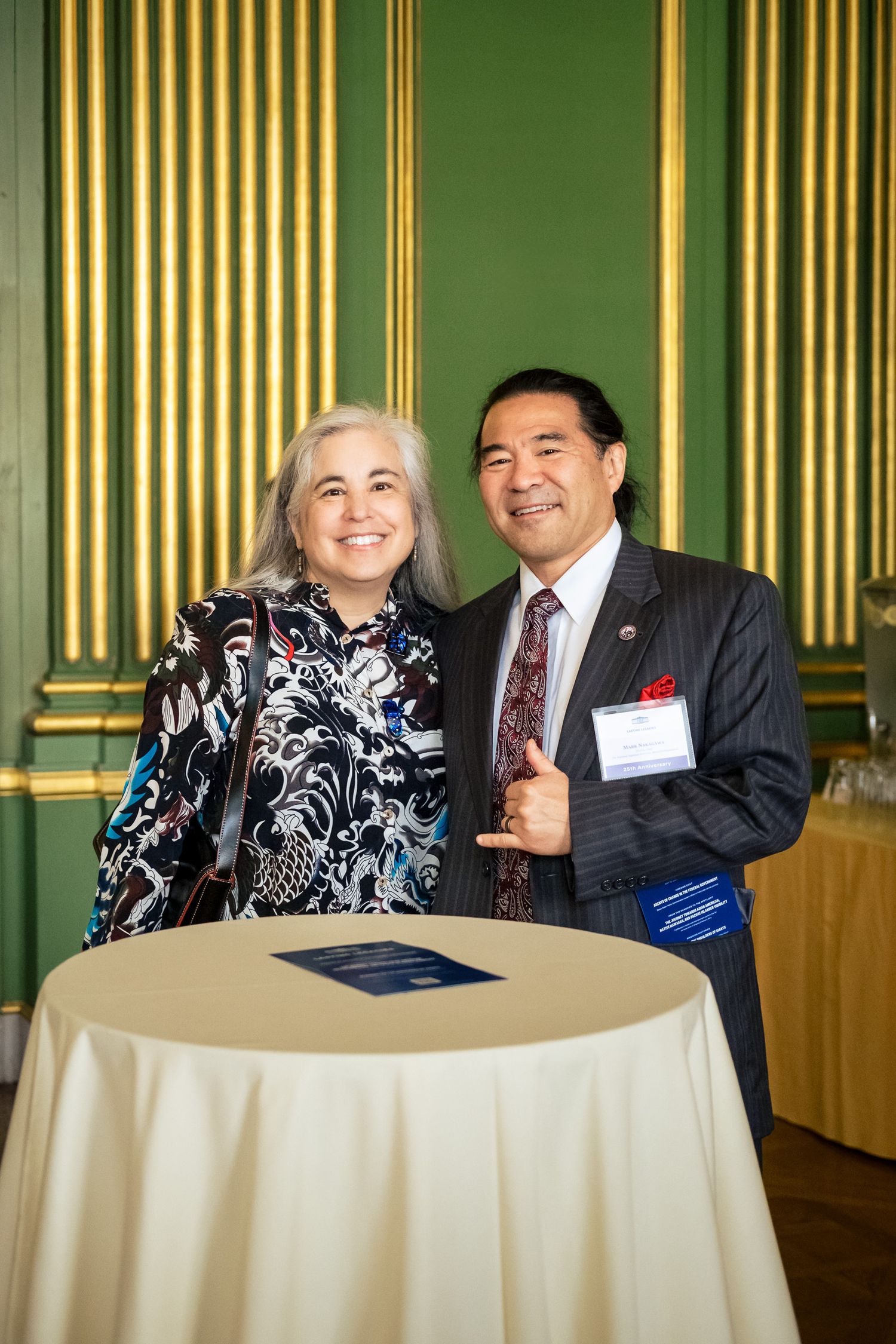 A man and woman smile together at a cocktail table in an elegant green and gold ballroom at a formal event.