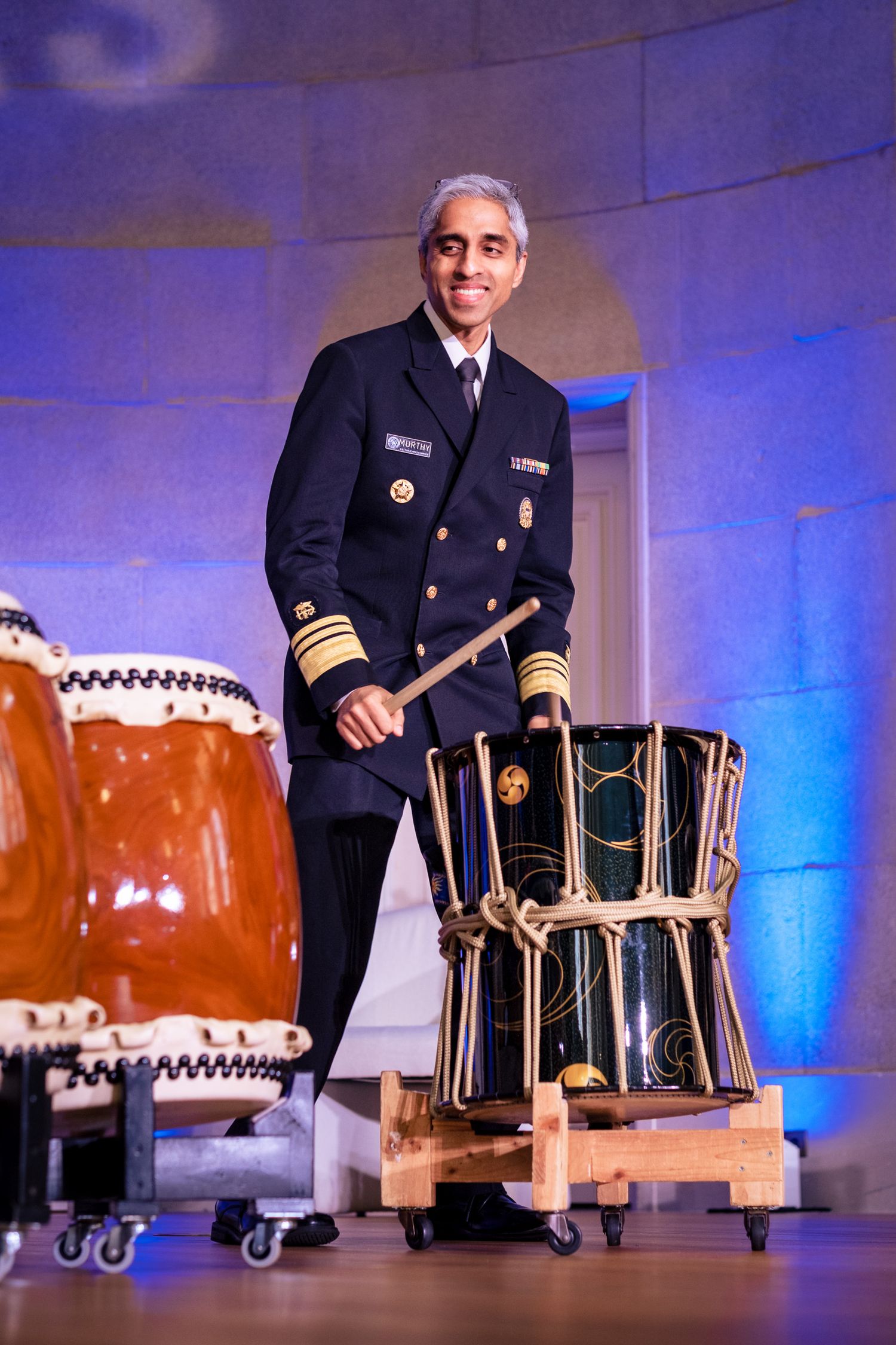 Military officer in navy uniform playing traditional taiko drums on a decorated stage with blue lighting.