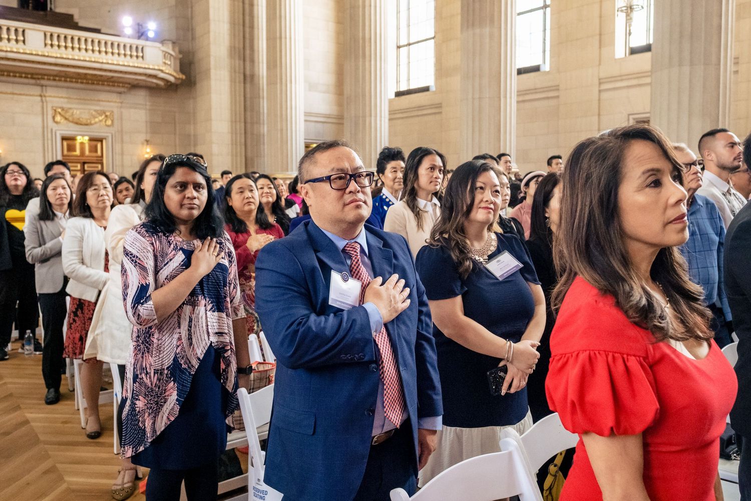 Diverse group of people standing with hands over hearts during a naturalization ceremony in a grand hall.