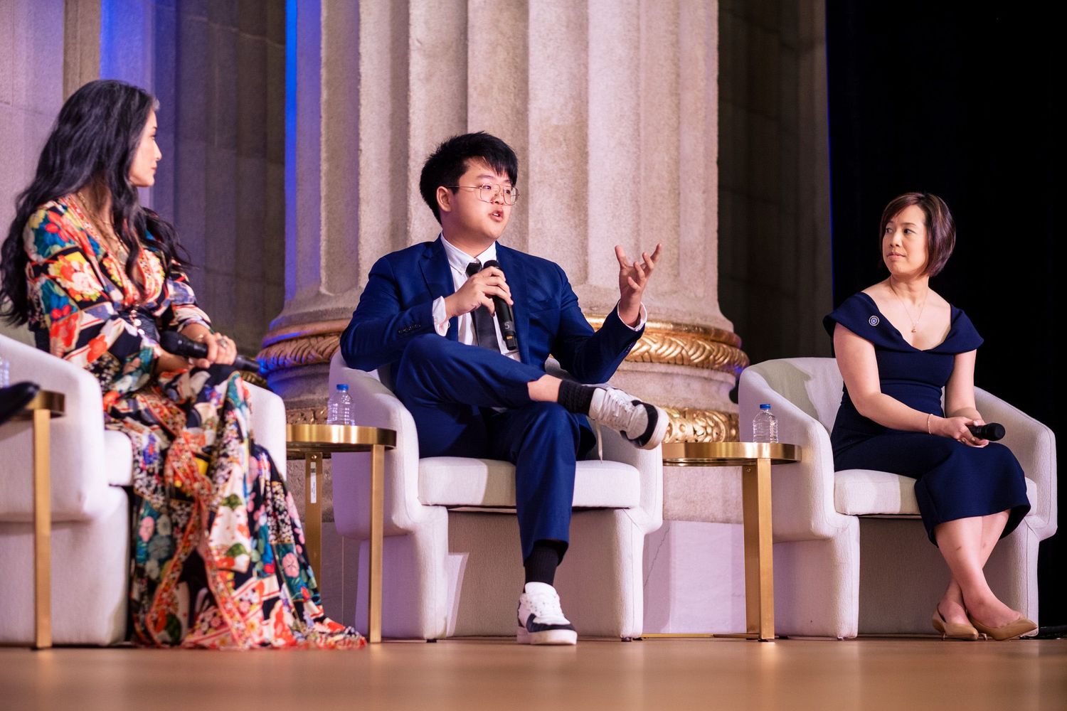 Three panelists seated on stage in elegant chairs during a formal conference event, with a man in blue suit gesturing.