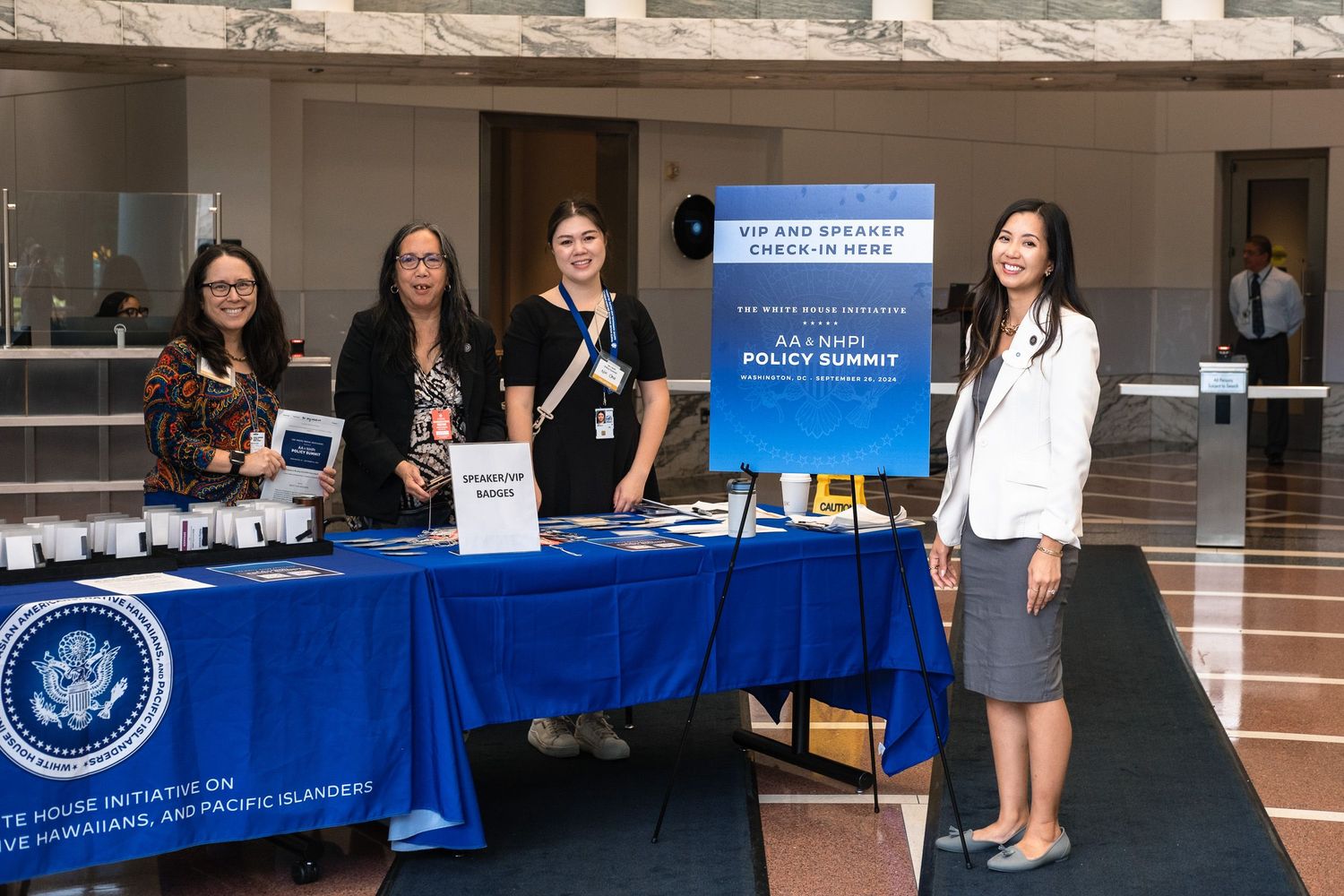 Four women stand at a registration table for an annual policy summit event in a government building.