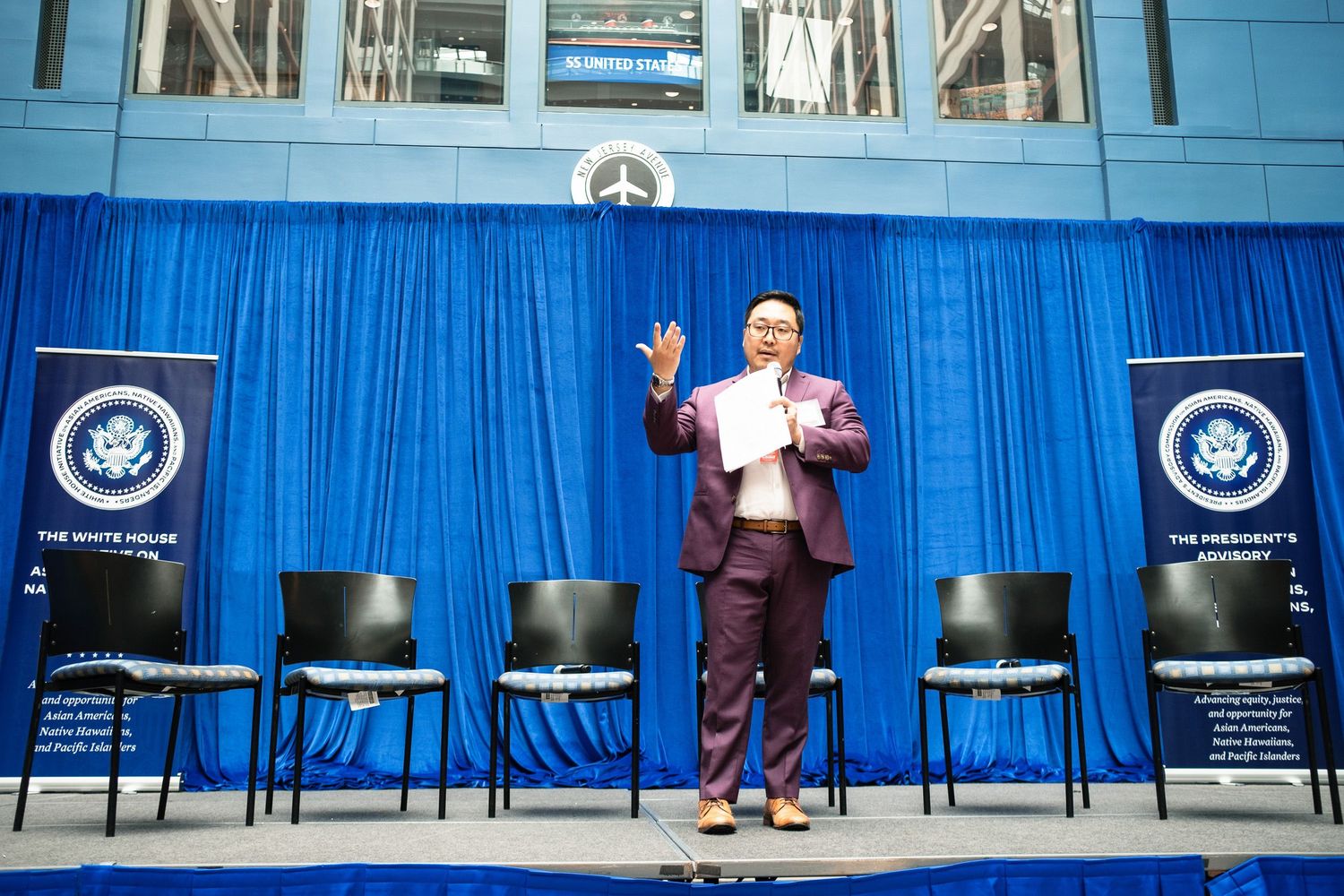 Speaker presenting at White House initiative event on stage with blue backdrop and presidential banners.