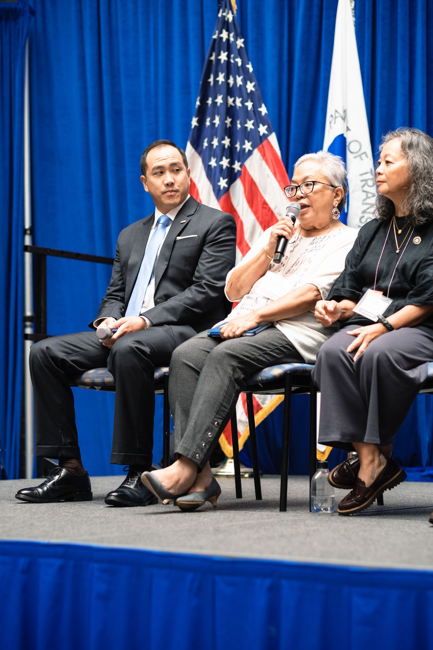 Three panelists seated on stage at a formal event with US flag backdrop, middle speaker holds microphone.