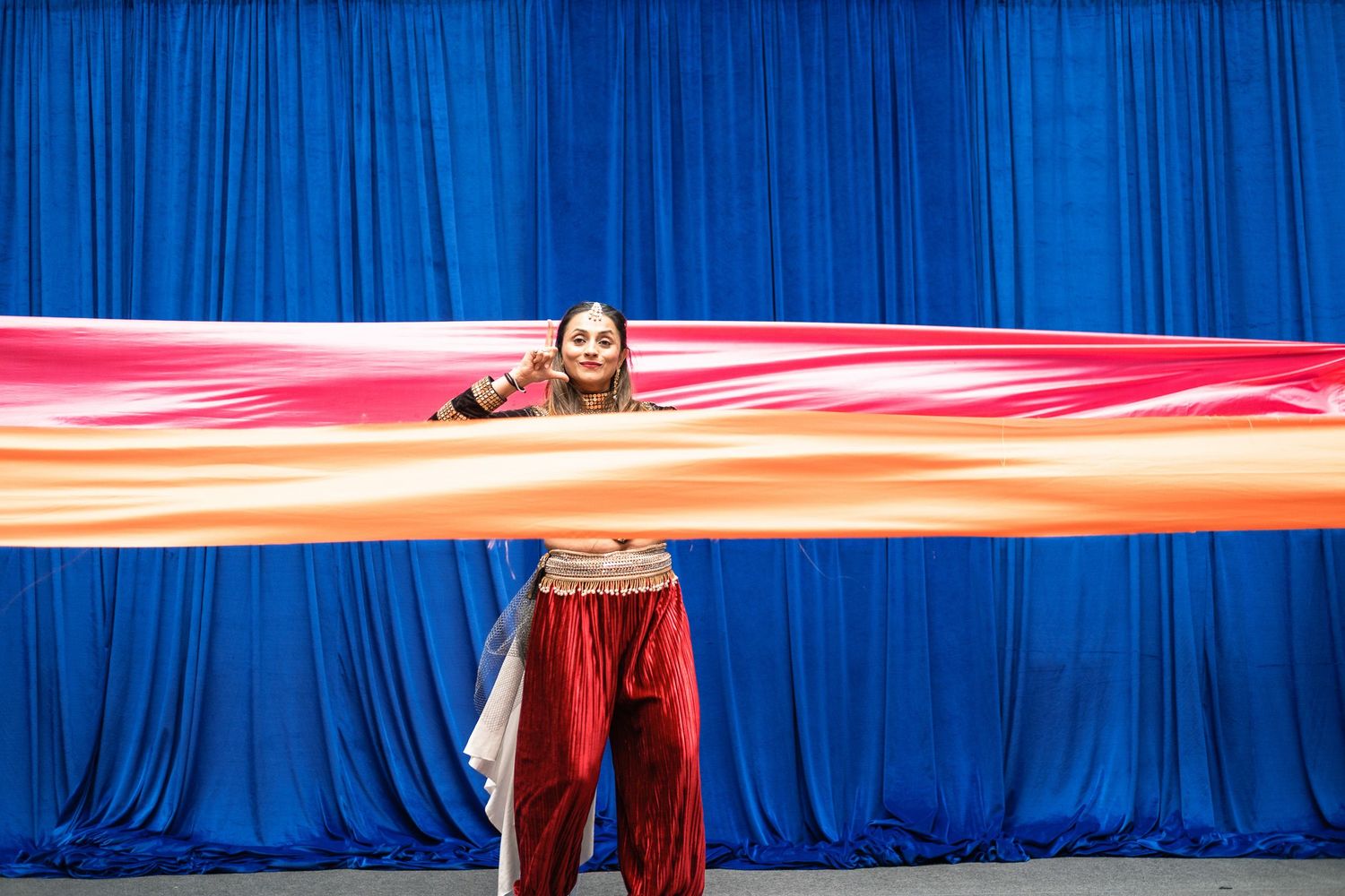 Belly dancer in red costume holds a large colorful silk veil on stage with blue curtain backdrop.