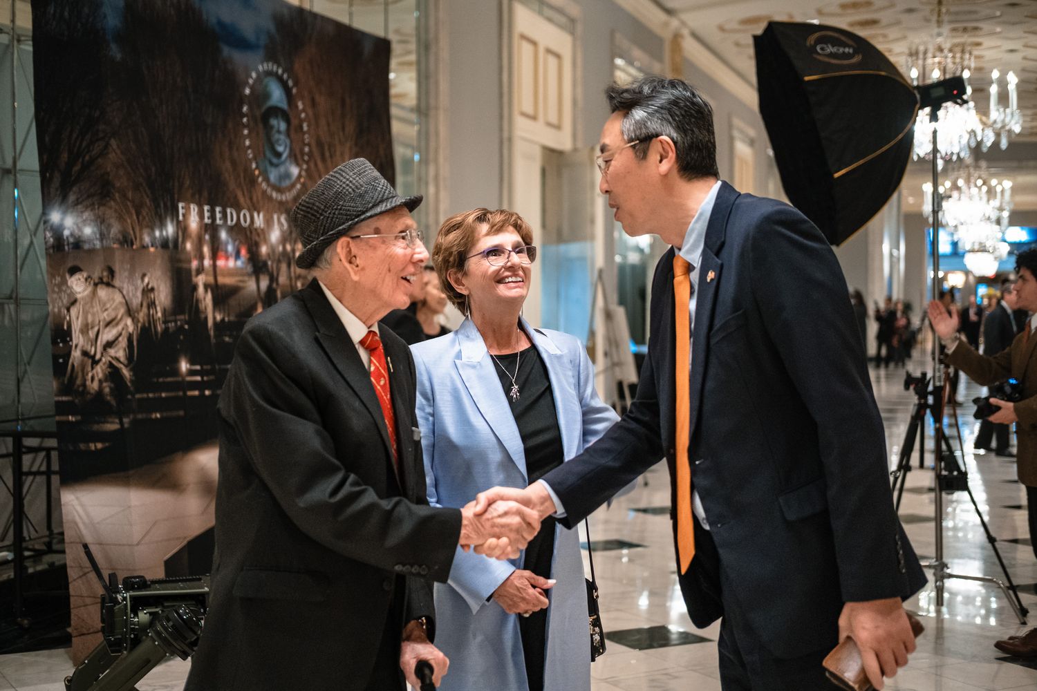 Three people shaking hands and smiling at a formal indoor event with elegant decor and photography lighting.