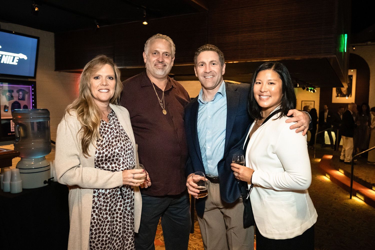 Four smiling adults pose together at an indoor social event in a dimly lit venue.