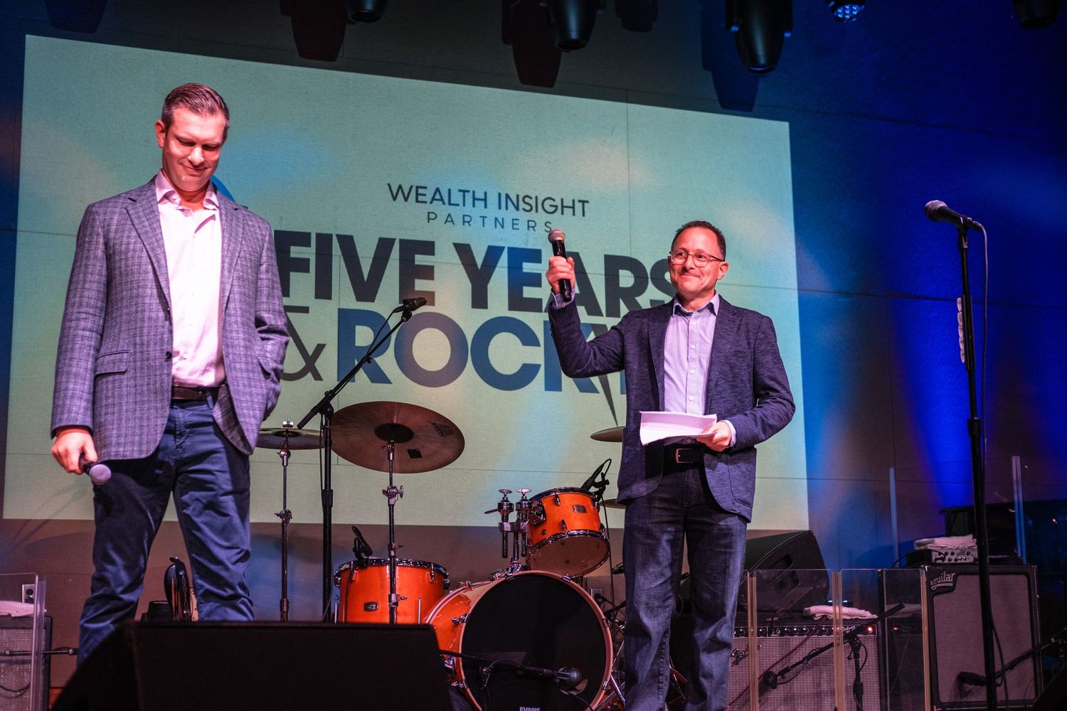 Two men on stage at Wealth Insight Partners Five Years Rock anniversary event with drum kit and microphone stand.