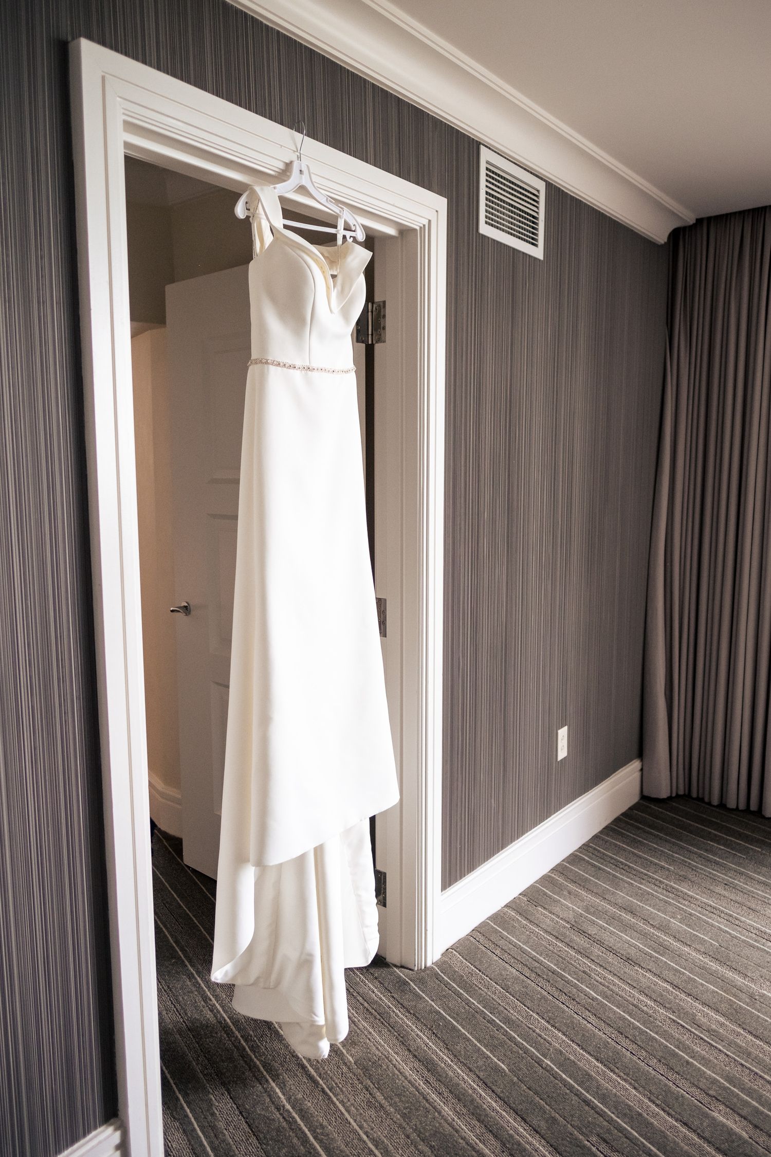 Elegant white sleeveless wedding gown hanging in hotel room doorway on wedding day.