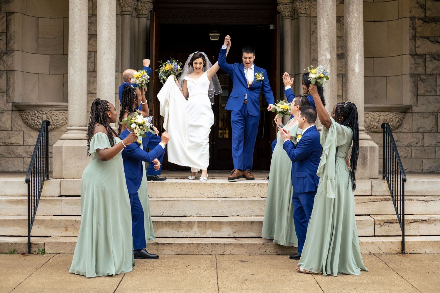 Joyful bride and groom raise hands while wedding party in sage and blue celebrate on church steps.