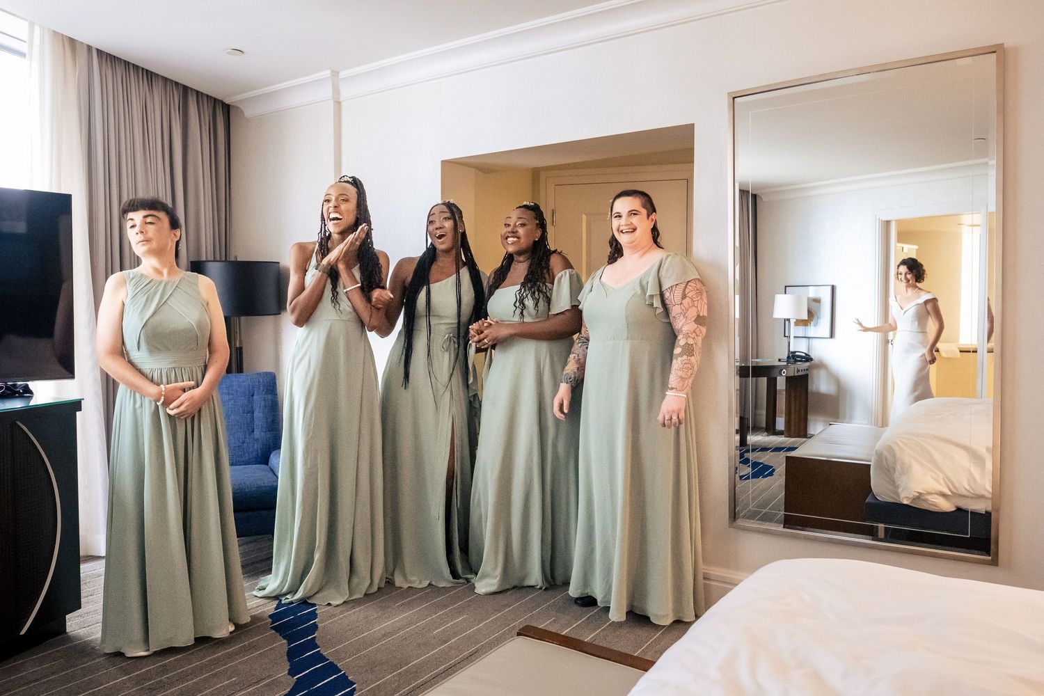 Four bridesmaids in sage green gowns react joyfully to bride during wedding morning preparations.