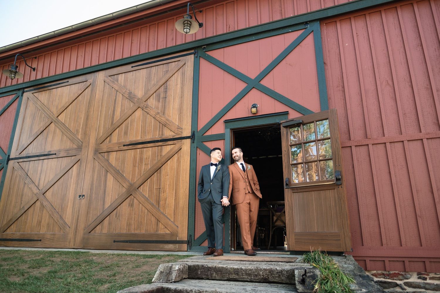 Two grooms in elegant suits stand at the entrance of a rustic red barn wedding venue.