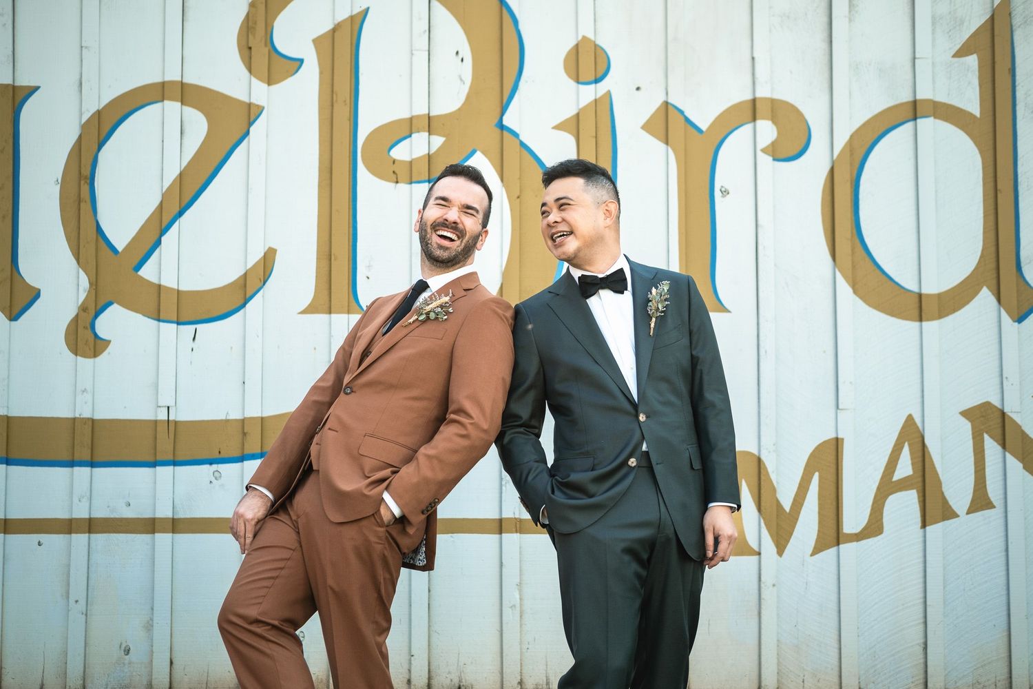 Two grooms laugh together in brown and dark teal suits at Blue Bird Manor.