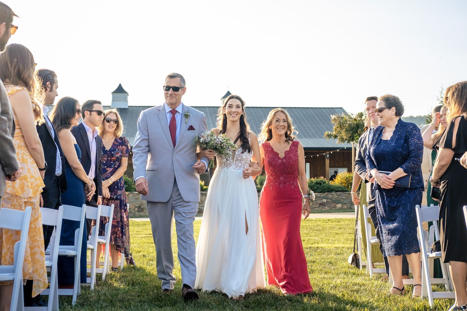 Bride walks down the aisle escorted by two family members during an outdoor wedding ceremony on a sunny day.
