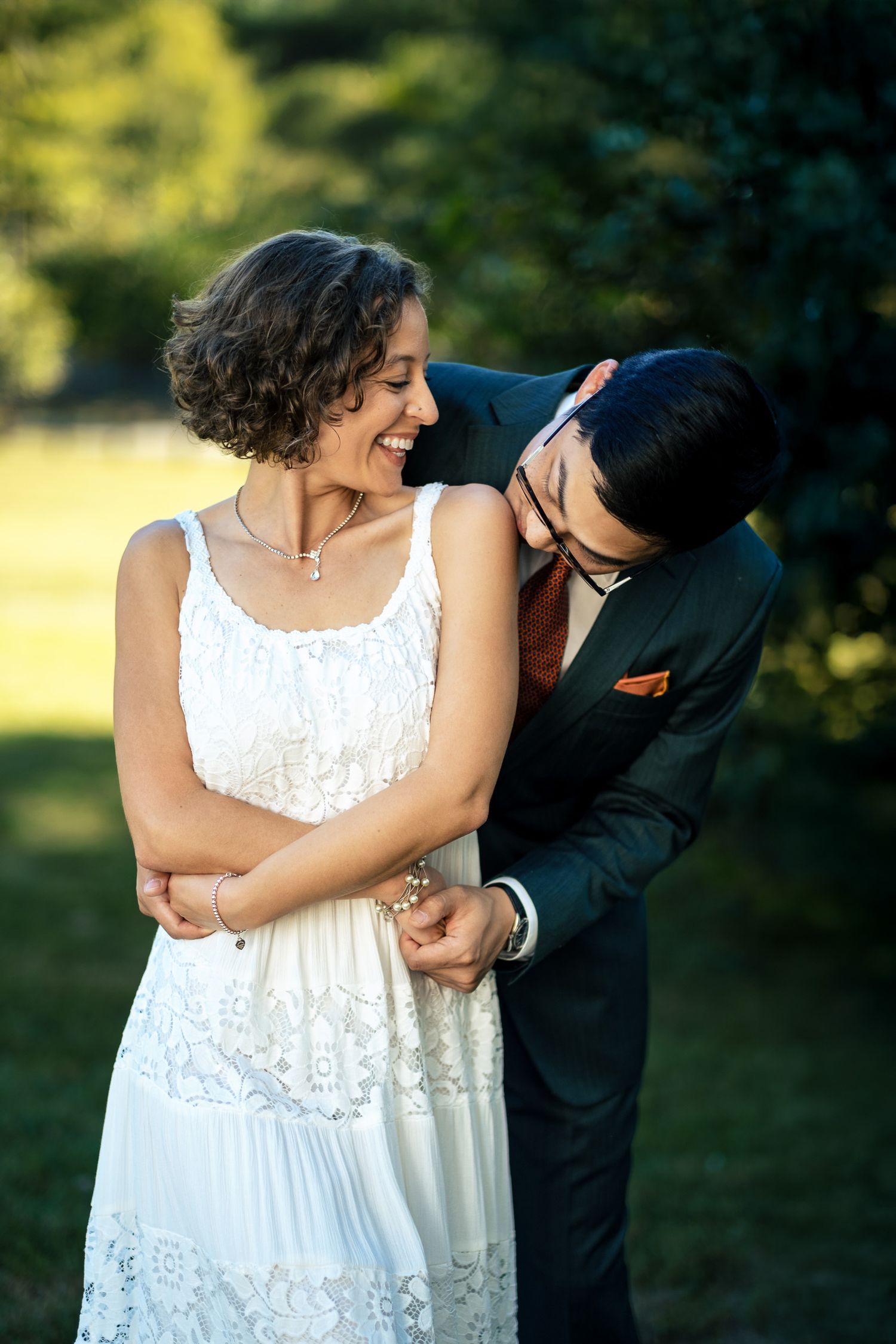 Groom in dark suit affectionately nuzzling laughing bride in white lace dress in a lush green garden.
